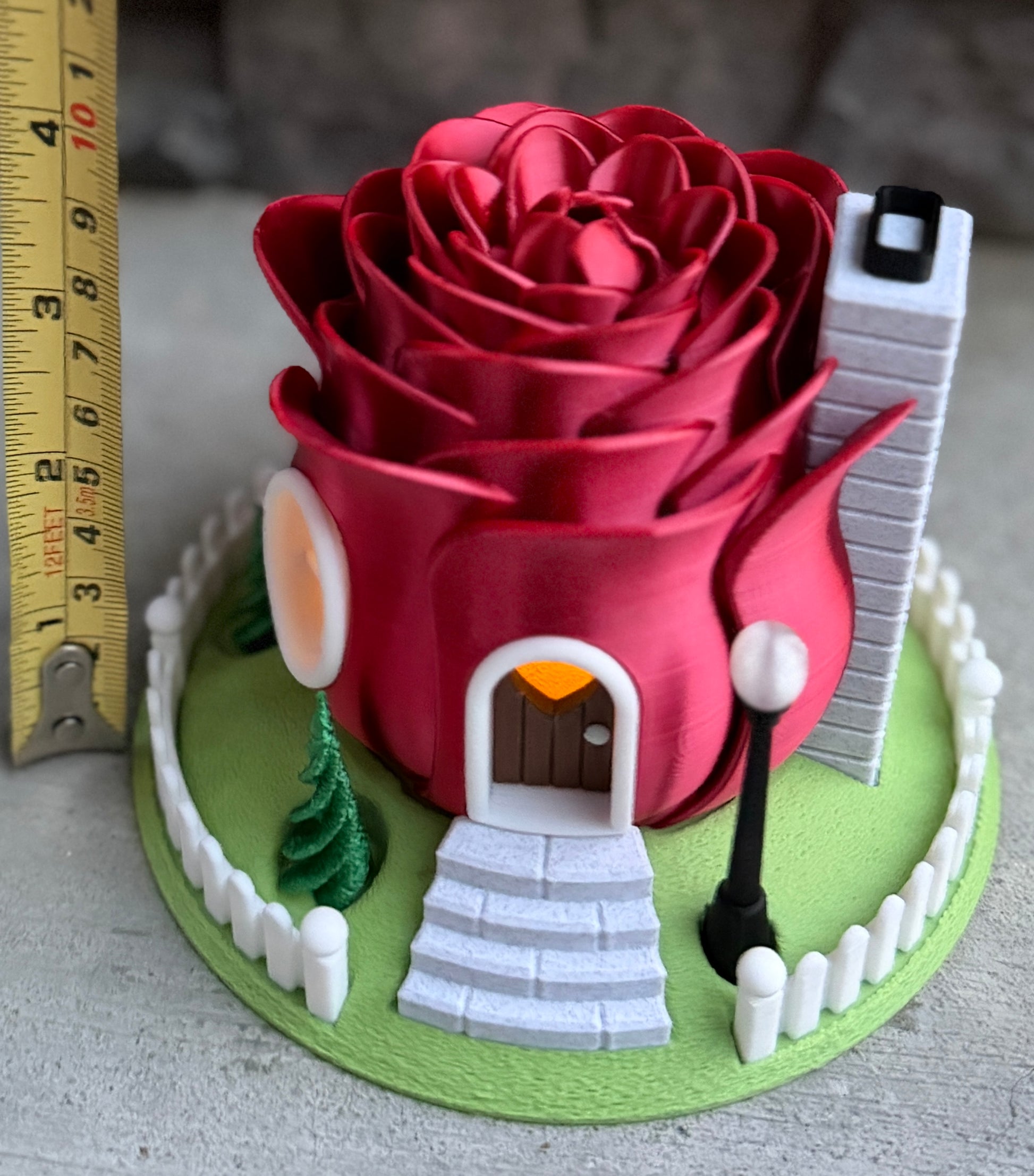 Red rose-shaped house with a door and windows on a green base, next to a ruler for scale.