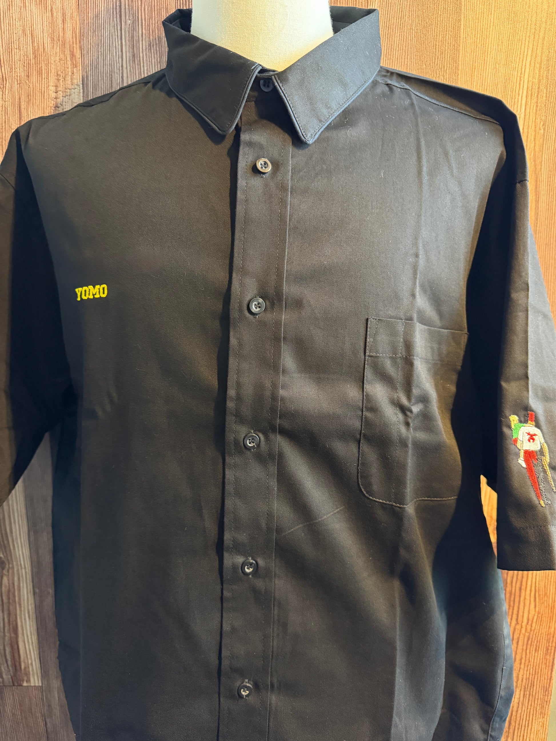 A dark charcoal gray button-up shirt with a pointed collar, displayed on a mannequin torso against a wood-paneled background. The shirt features a yellow embroidered 'YOMO' text on the left chest and a small colorful graphic on the right sleeve.