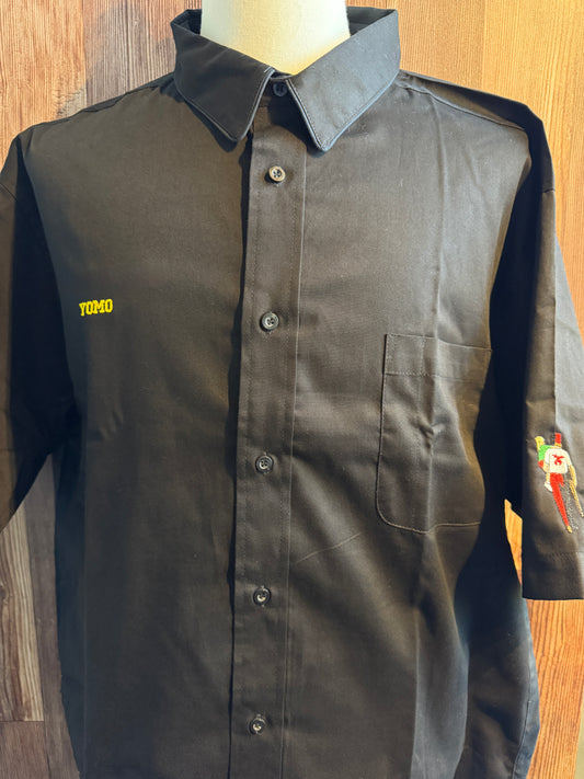 A dark charcoal gray button-up shirt with a pointed collar, displayed on a mannequin torso against a wood-paneled background. The shirt features a yellow embroidered 'YOMO' text on the left chest and a small colorful graphic on the right sleeve.