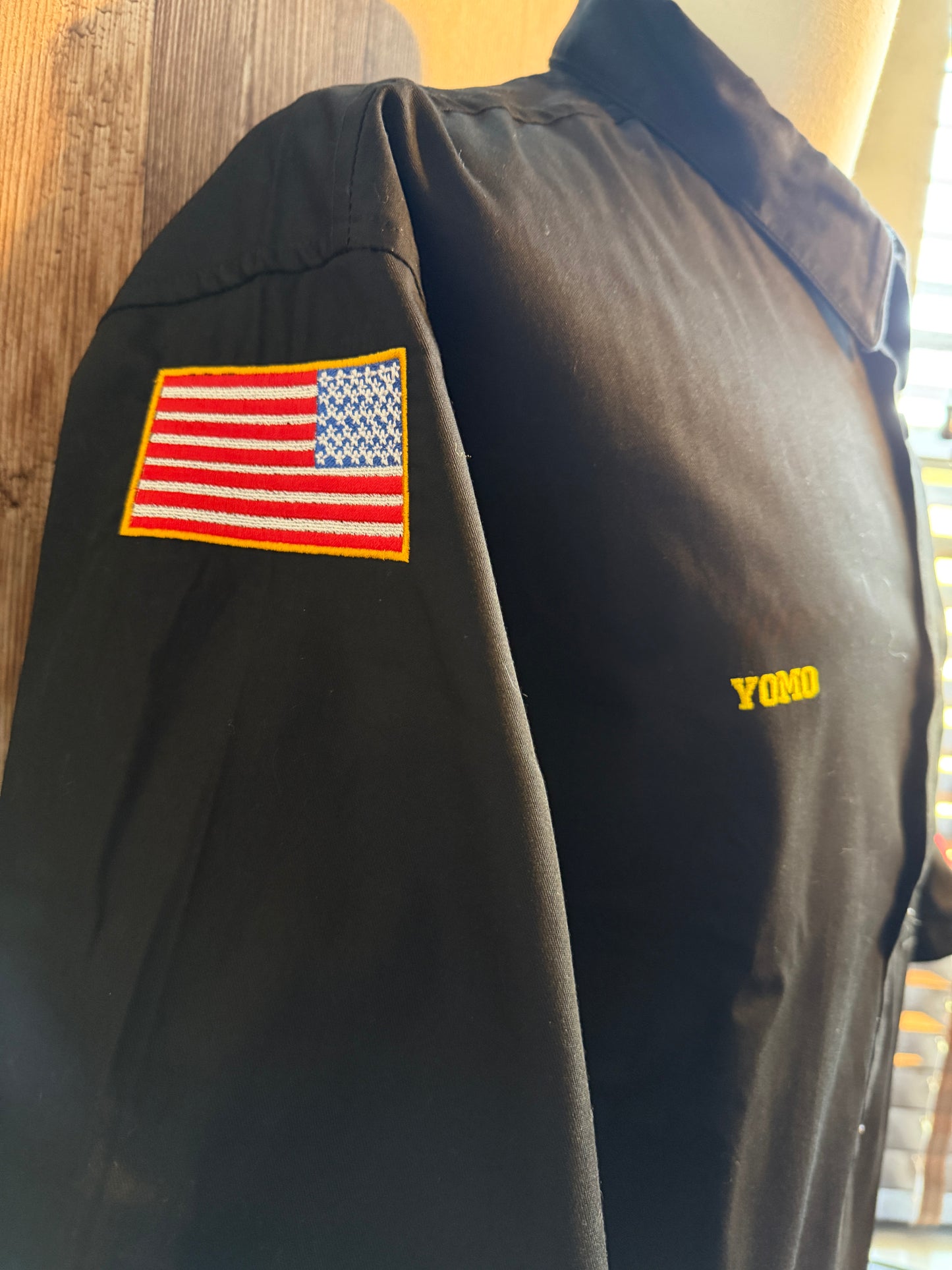 A black work shirt displayed on a mannequin torso, featuring a yellow embroidered 'YOMO' logo on the chest and an embroidered American flag patch on the right sleeve.