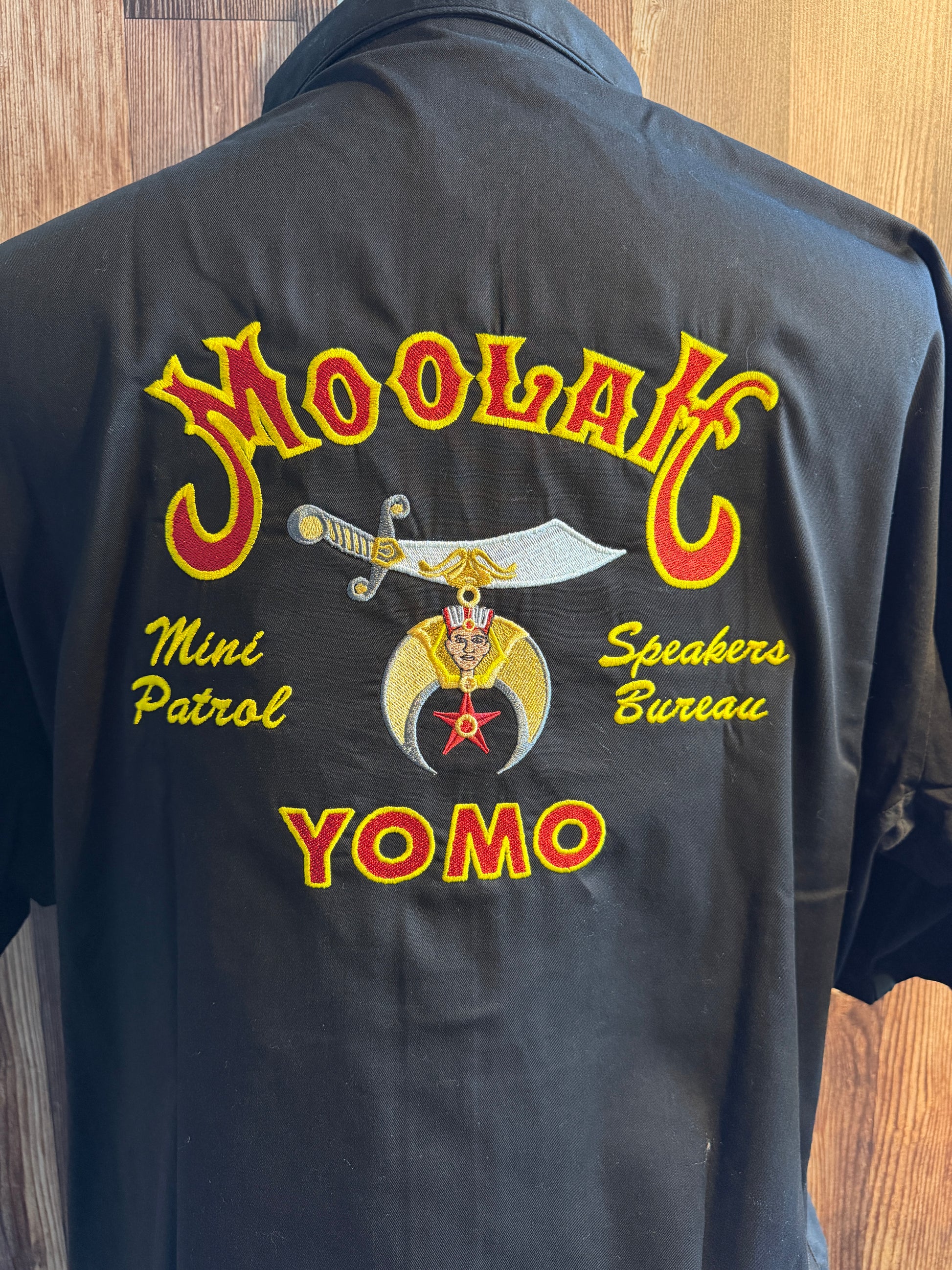 A black short-sleeve button-up shirt shown from the back, featuring a large embroidered graphic with the words 'MOOLAH, 'Mini Patrol', 'Speakers Bureau', and 'YOMO' in yellow and red, plus a central emblem with a sword and star.